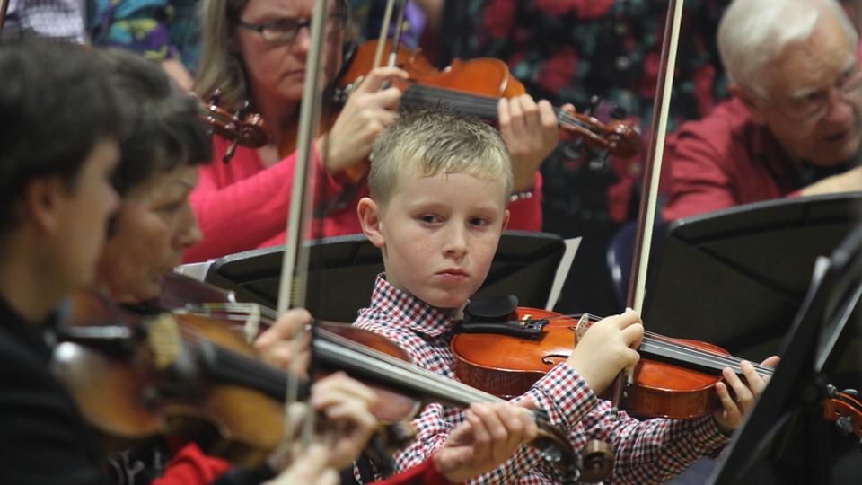 A young boy holds a violin and watches other violinists play