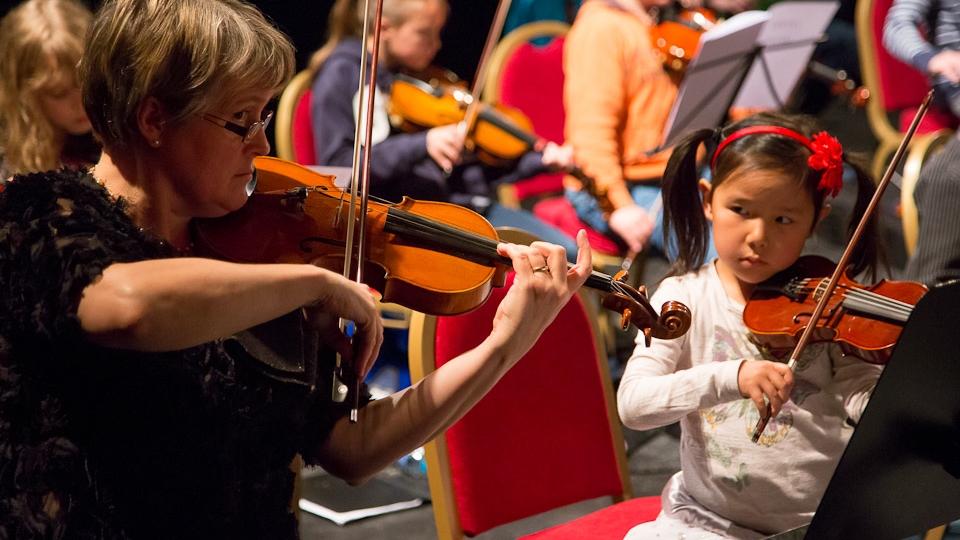 A young girl is playing a violin as she watches an older woman play