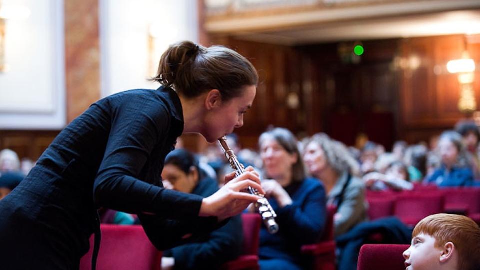 A woman plays flute to a young boy in an audience 